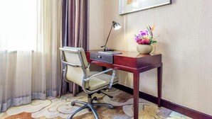 A hotel room with a desk, chair, lamp, and a framed picture on the wall.