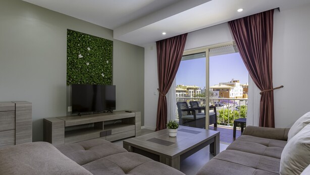 A modern living room with a flat-screen TV, a coffee table, and a sofa. There is a balcony with a view of buildings and a railing. - Corcovada Sunset (Albufeira)