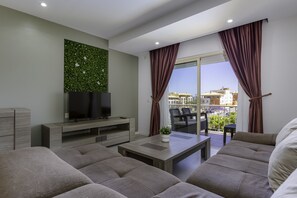 Ein modernes Wohnzimmer mit Flachbildfernseher, Couchtisch und Sofa. Ein Balkon mit Blick auf Gebäude und Geländer ist vorhanden.