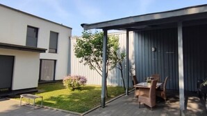 Ein überdachter Patio-Bereich mit Tisch und Stühlen, eine kleine Rasenfläche und ein Gebäude im Hintergrund.