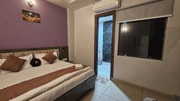 A bedroom with a bed, a nightstand, an air conditioning unit, a window, and a door leading to a bathroom.