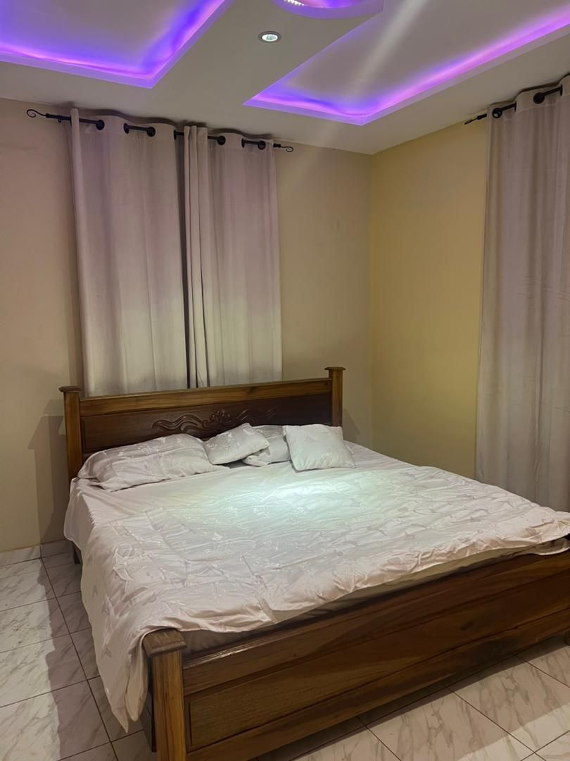 Una cama bien tendida en una habitación con cabecera de madera, cortinas blancas e iluminación LED morada en el techo.