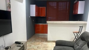 Una sala de estar con un sofá gris, un televisor, un escritorio de madera y una zona de cocina con armarios rojos y una ventana con persianas.