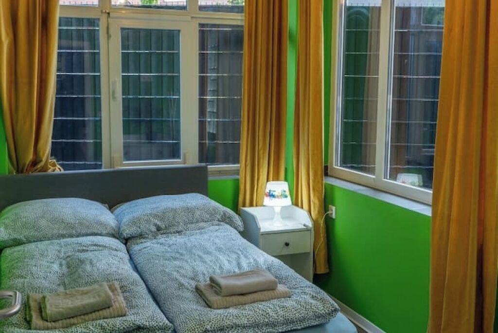 A bedroom with a bed, two pillows, a nightstand with a lamp, and a window with curtains.
