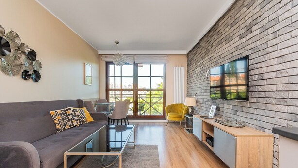 A modern living room with a grey sofa, a glass coffee table, and a yellow armchair. A TV is mounted on a brick wall, and there is a large window with a view of greenery. - Jaglana Deluxe 2 by Grand Apartments (Gdansk)