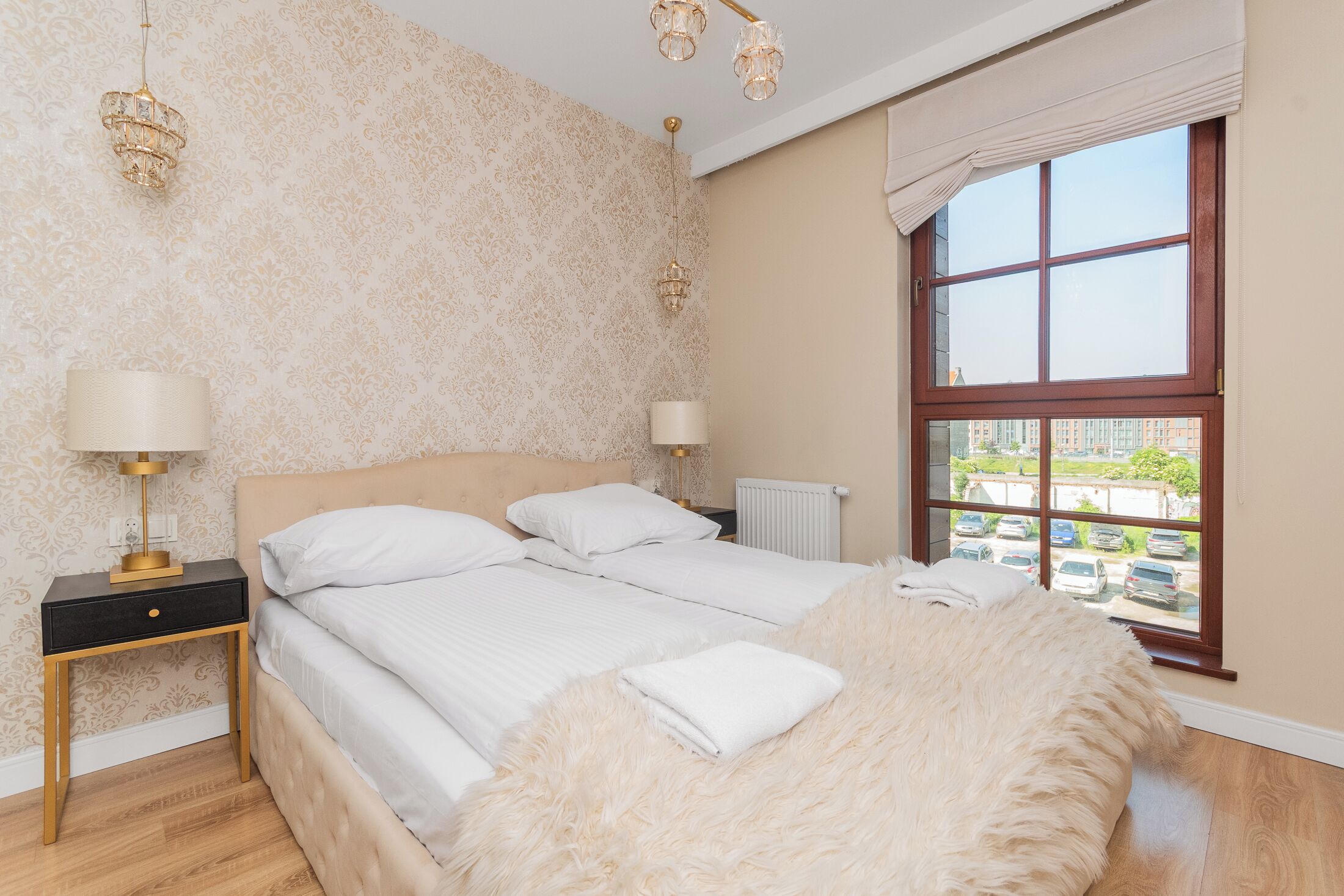 A bedroom with a large bed, two bedside lamps, a nightstand, a fluffy white throw, and a window with a view of parked cars and buildings.