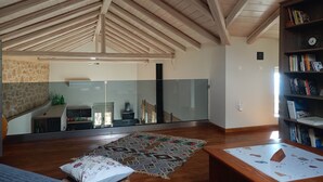 A spacious living area with a high wooden ceiling, a large glass door leading to a balcony, a wooden bookshelf, and a patterned rug on the floor.