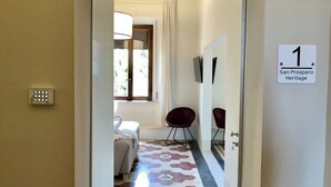 A hallway with a geometric patterned floor, a bed with a swan-shaped pillow, a red chair, and a sign indicating room number 1. - San Prospero Heritage (Siena)
