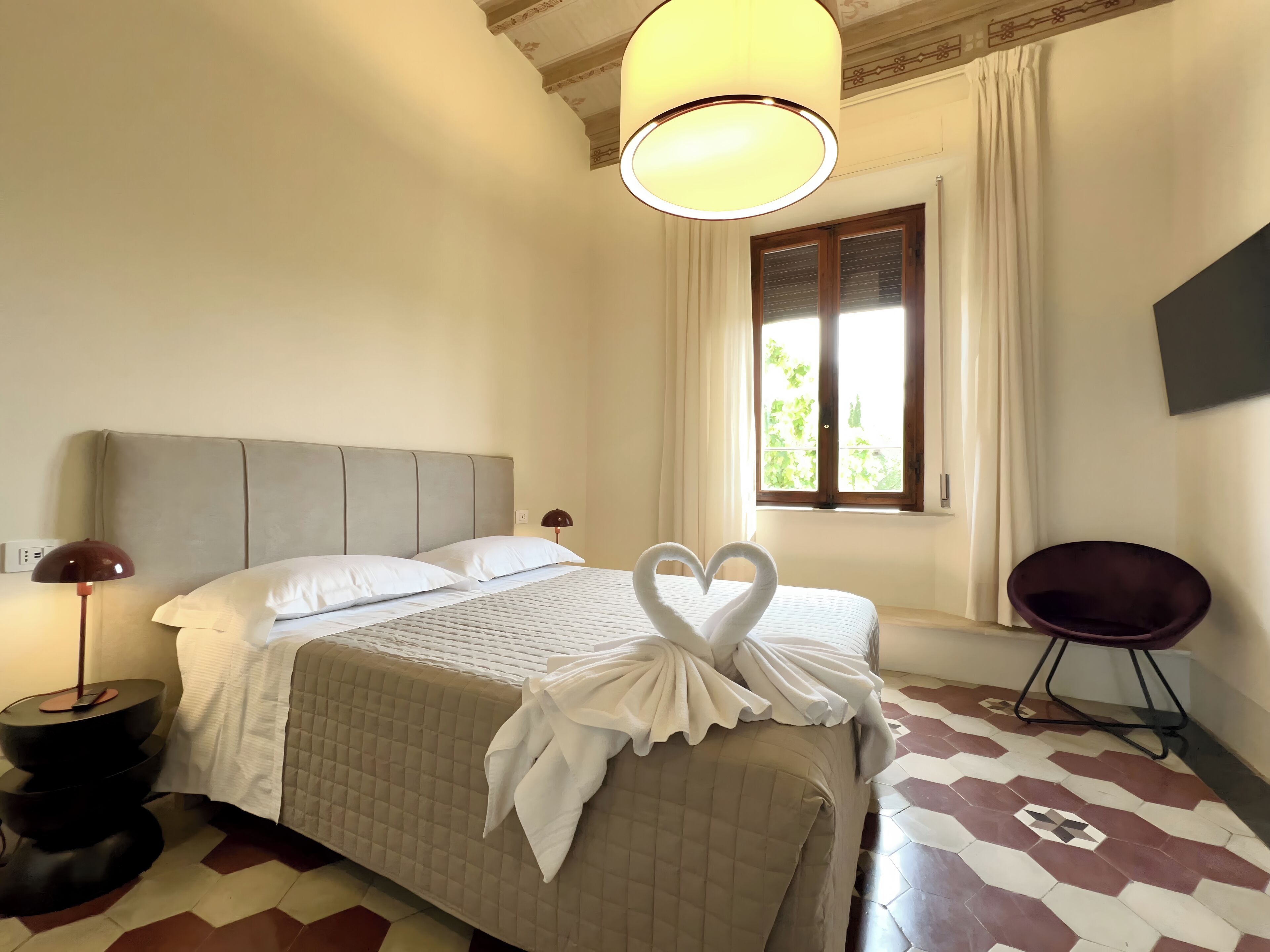 A hotel room with a bed, a window with curtains, a wall-mounted TV, and a chair.