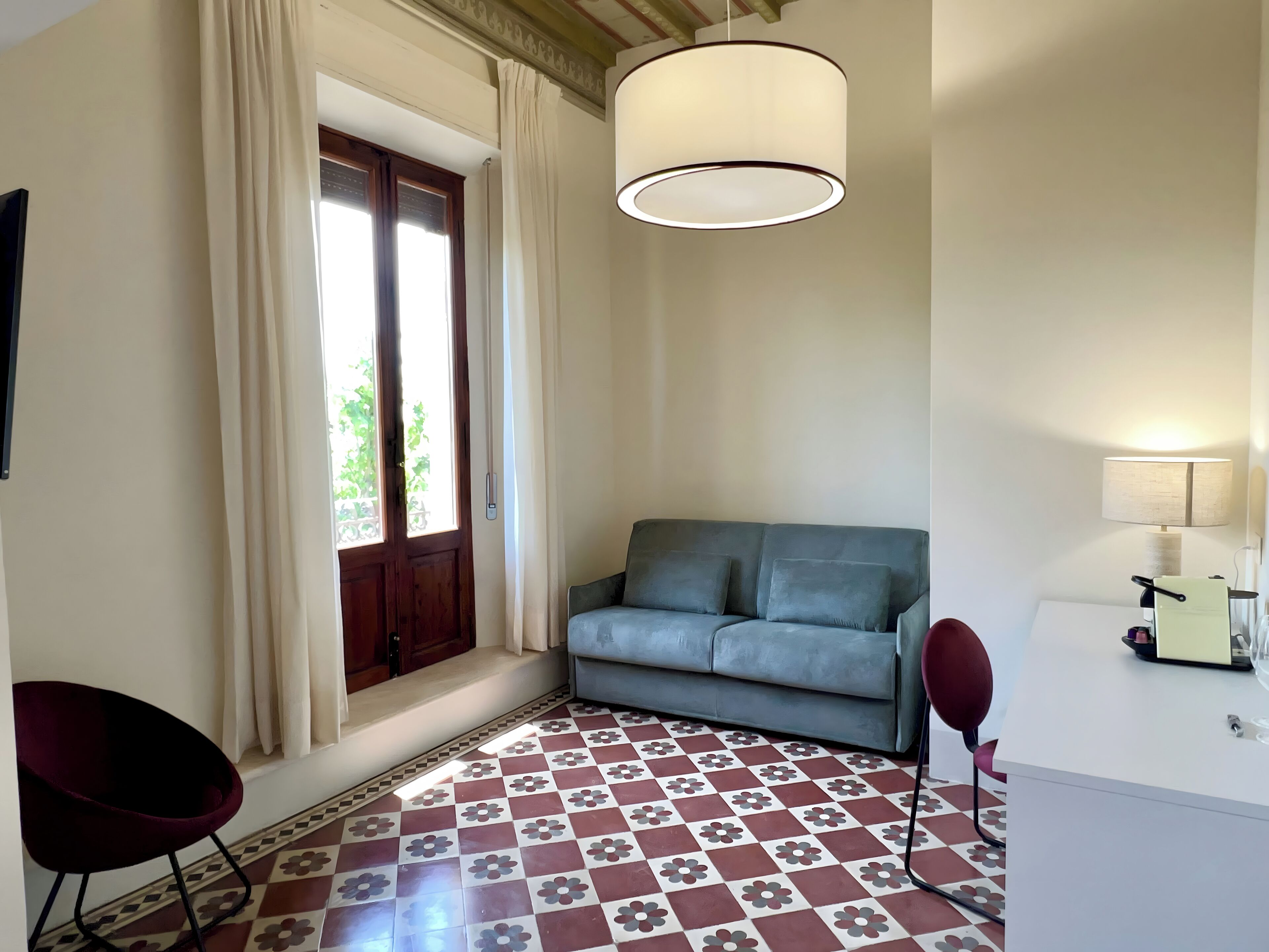 Ein Zimmer mit Schachbrettmuster auf dem Boden, einem grauen Sofa, einem roten Sessel, einem weißen Schreibtisch und einer hängenden Lampe.
