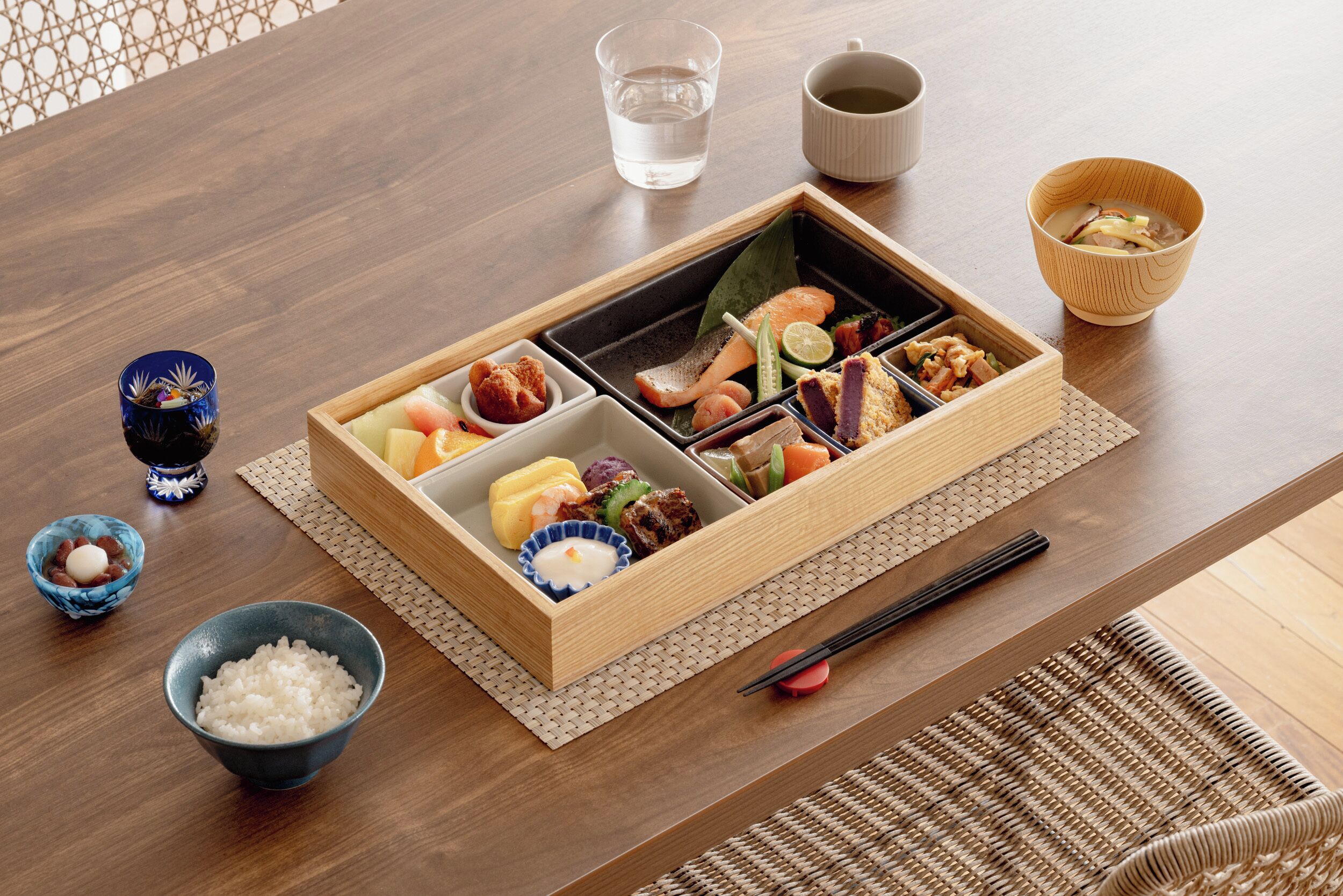 A wooden tray with various food items, a glass of water, a cup, and a bowl on a table.