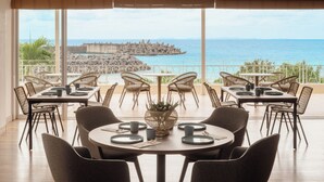 Een eethoek met tafels en stoelen, een groot raam met uitzicht op zee en rotsen, en een balkon met rieten meubels.