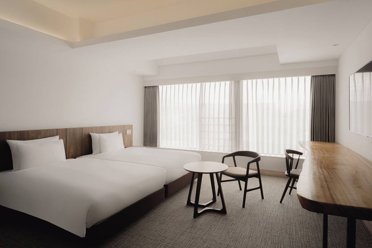 A hotel room with a large bed, a small table, two chairs, and a large window with curtains.