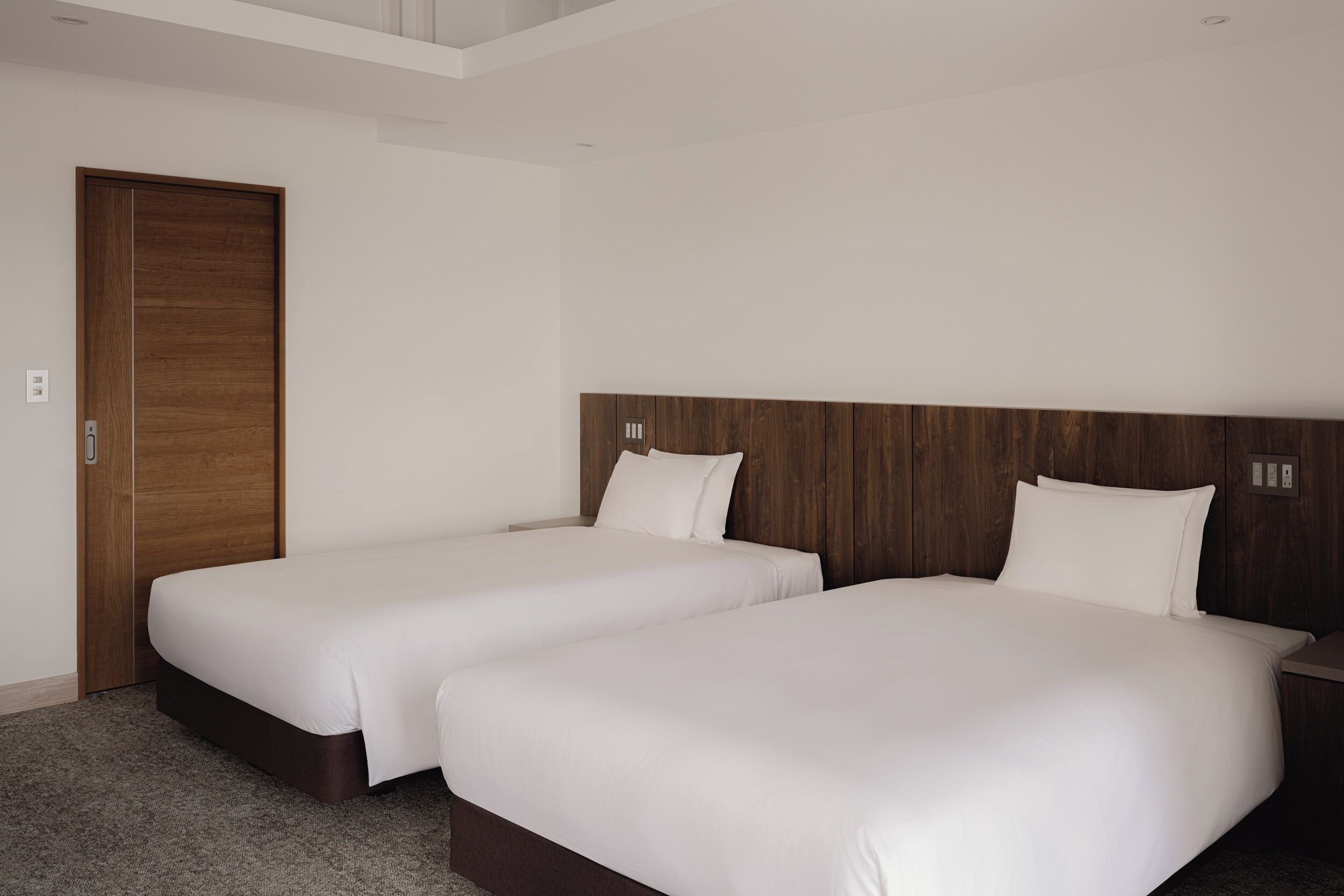 A hotel room with two single beds, a wooden headboard, a carpeted floor, and a door.
