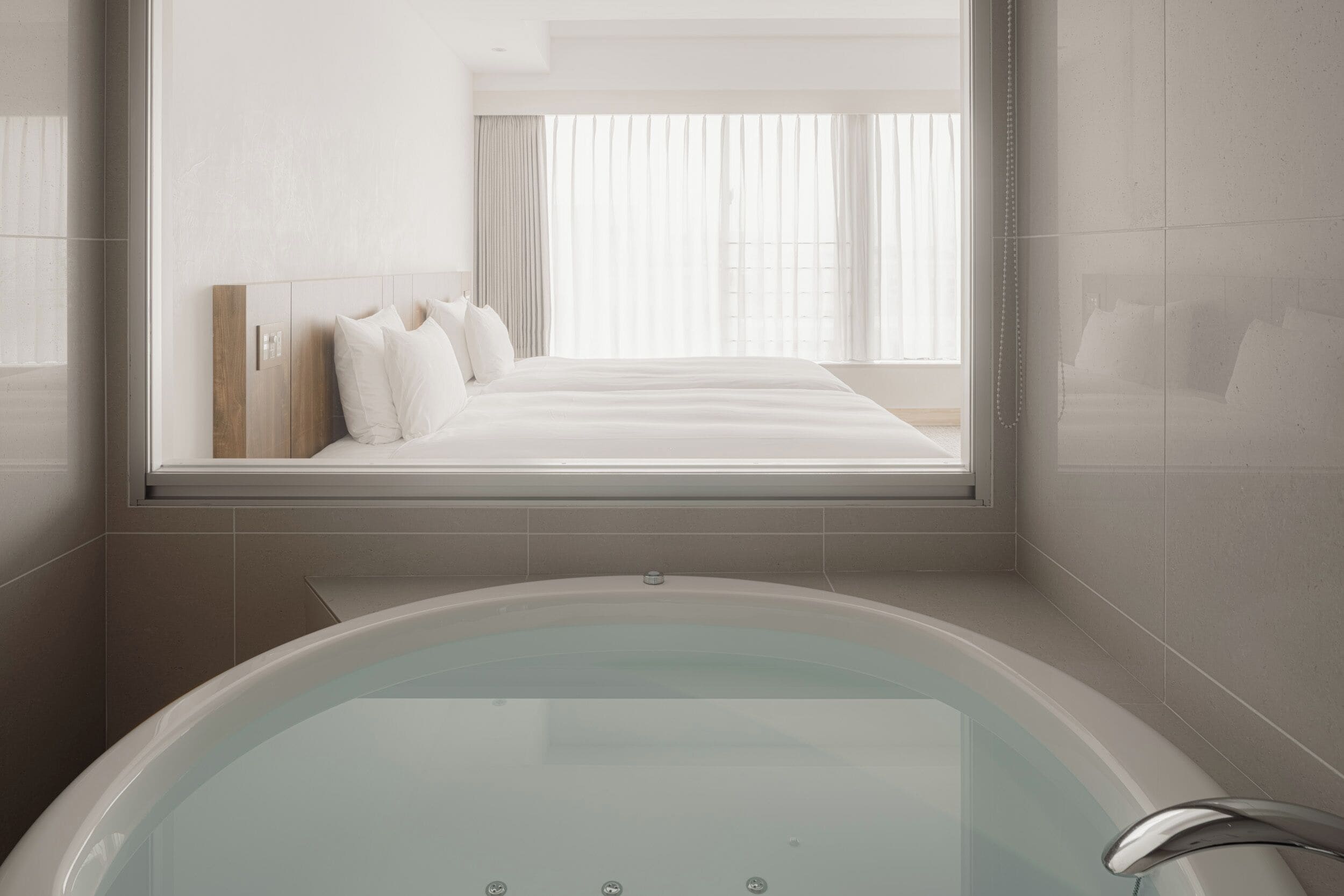 A modern bathroom with a large bathtub, a view of a bedroom through a window, and a mirror reflecting the room.