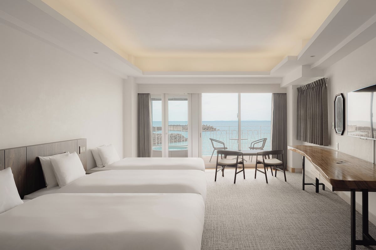 A hotel room with two beds, a desk, and a view of the sea.