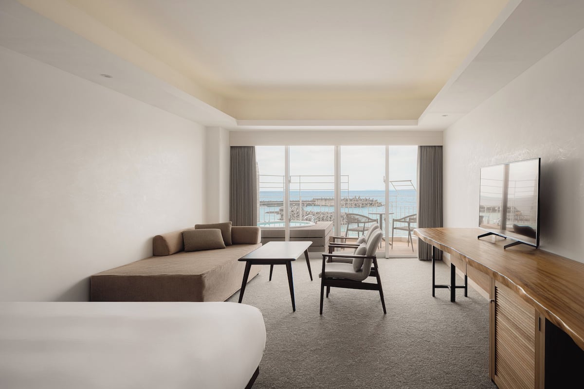 A hotel room with a large bed, a desk, a chair, a sofa, and a TV. There is a large window with a view of the sea.