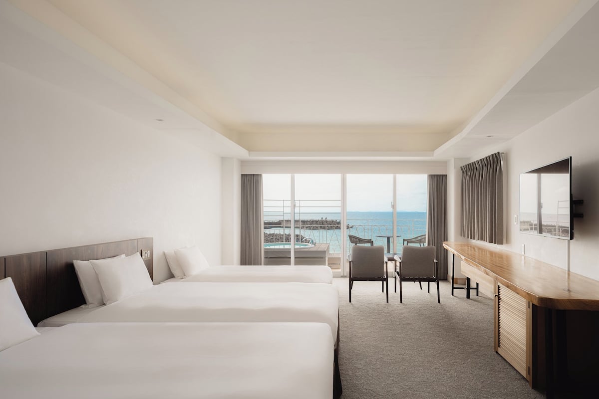 A hotel room with two beds, a desk, a chair, and a large window with a view of the sea.