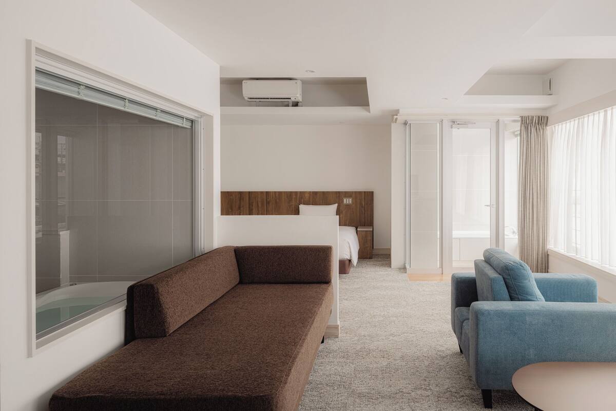 A modern hotel room with a large window, a sofa, a chair, a bed, and a shower area.