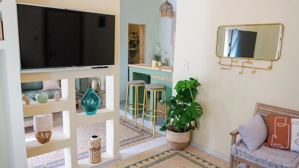 Ein Wohnzimmer mit Fernseher, Barhocker, einer großen Topfpflanze und einem Spiegel an der Wand.