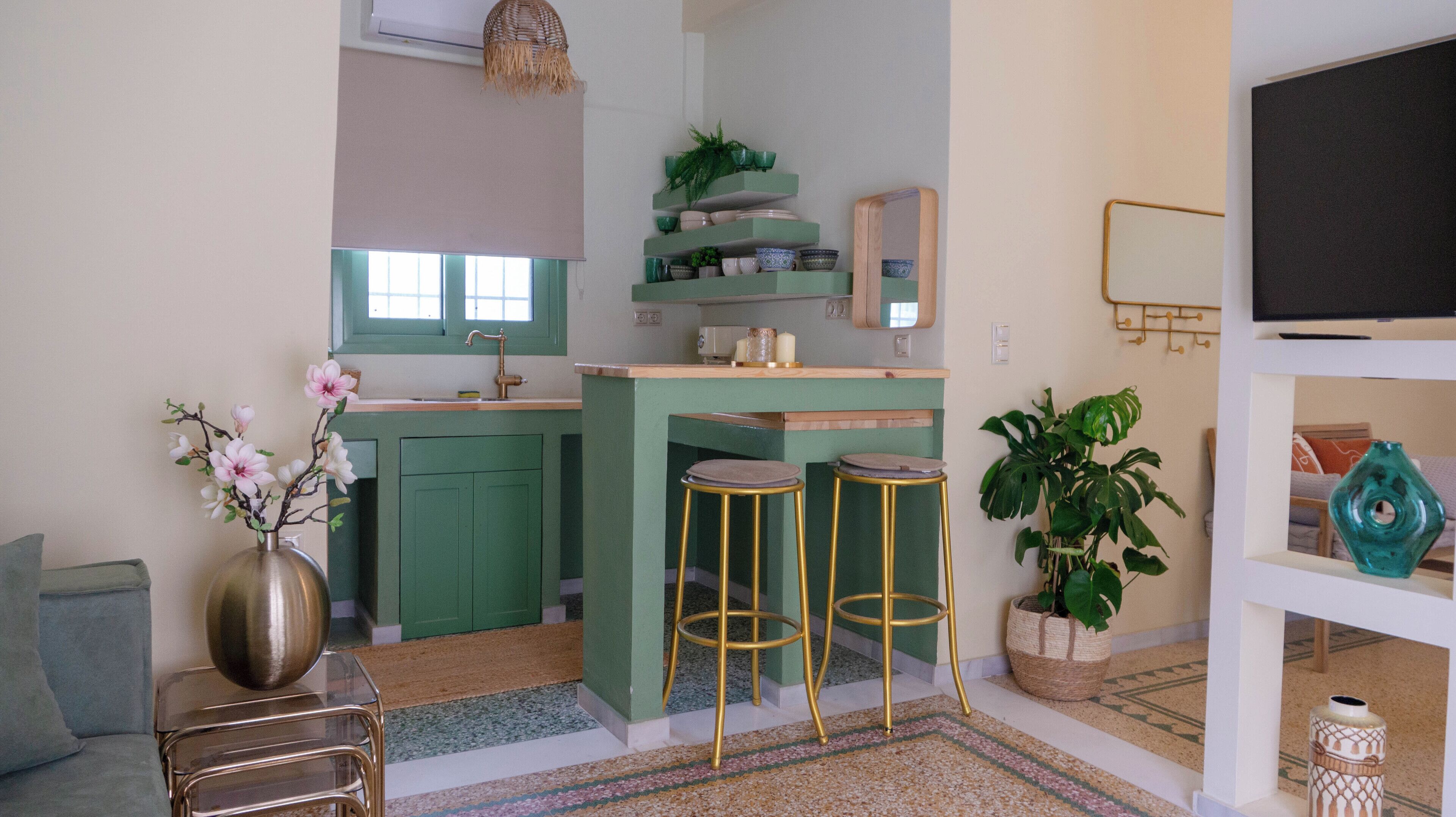 Cocina compacta con gabinetes verdes, un lavamanos y taburetes. Hay una planta en maceta y un florero con flores en la encimera.