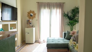 A compact living space with a kitchenette, a small refrigerator, a sofa, a television, and a large window with curtains. - Loft w/Rooftop in Athens – Spacious & Peaceful (Athina)