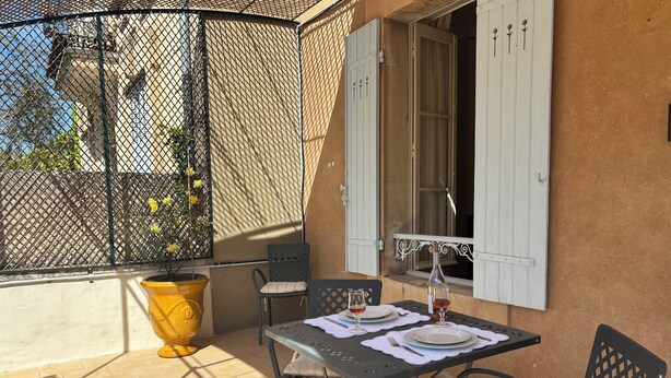 An outdoor patio with a table set for two, a potted plant, and a window with shutters.