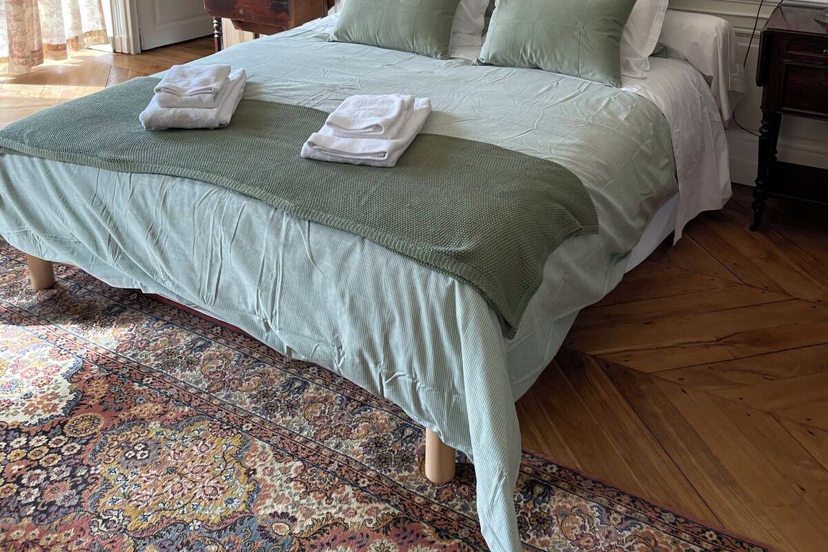 Uma cama bem arrumada com colcha e almofadas verdes, um tapete estampado e um quadro na parede.