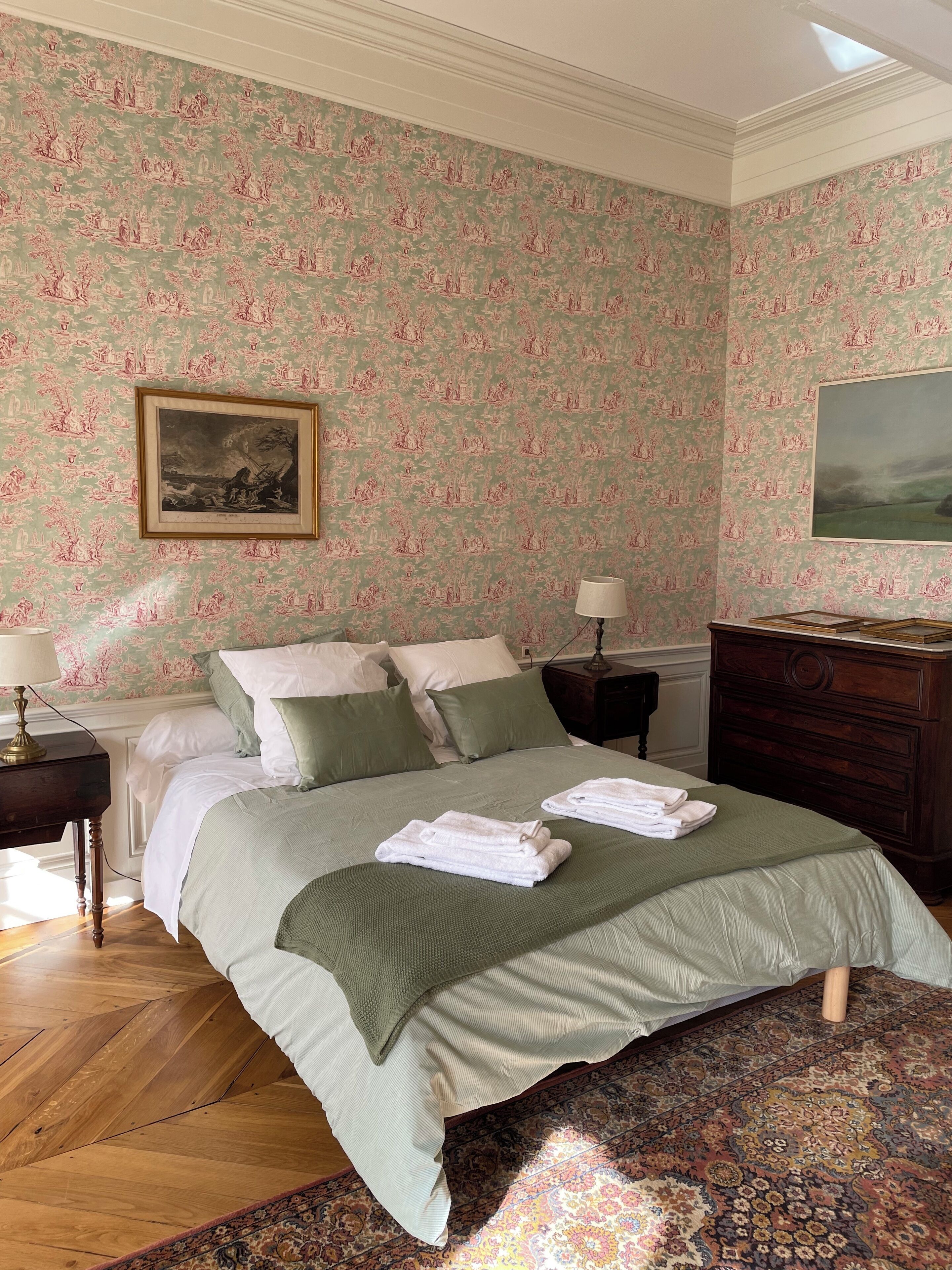 Een opgemaakt bed met een groene spreidek, twee kussens en opgevouwen handdoeken op een kamerkleed met dessin.