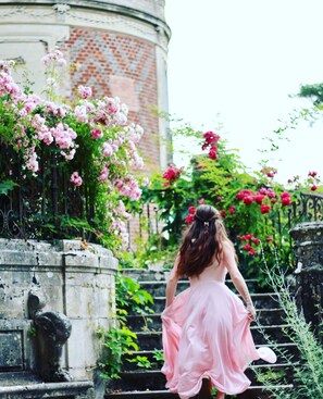 Eine Frau in einem rosafarbenen Kleid, die eine Steintreppe hinabsteigt, umgeben von blühenden rosafarbenen und roten Blumen.