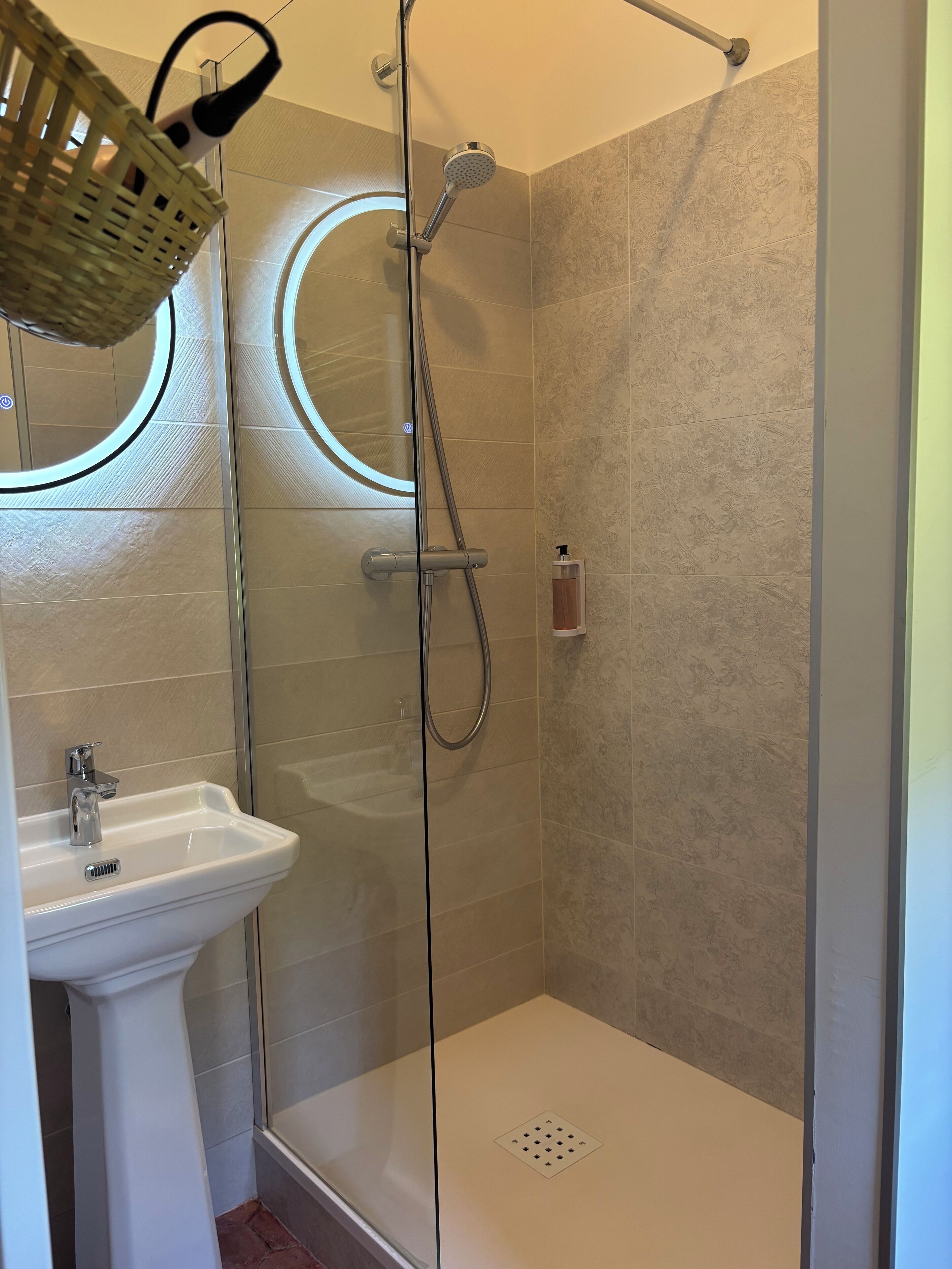 Un baño moderno con ducha envuelta en vidrio, un lavamanos pedestal blanco y un dispensador de jabón montado en la pared.