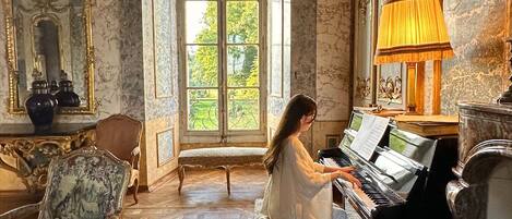 Une vaste pièce agrémentée d’un piano, de meubles richement décorés et d’une grande fenêtre offrant une vue sur la verdure.