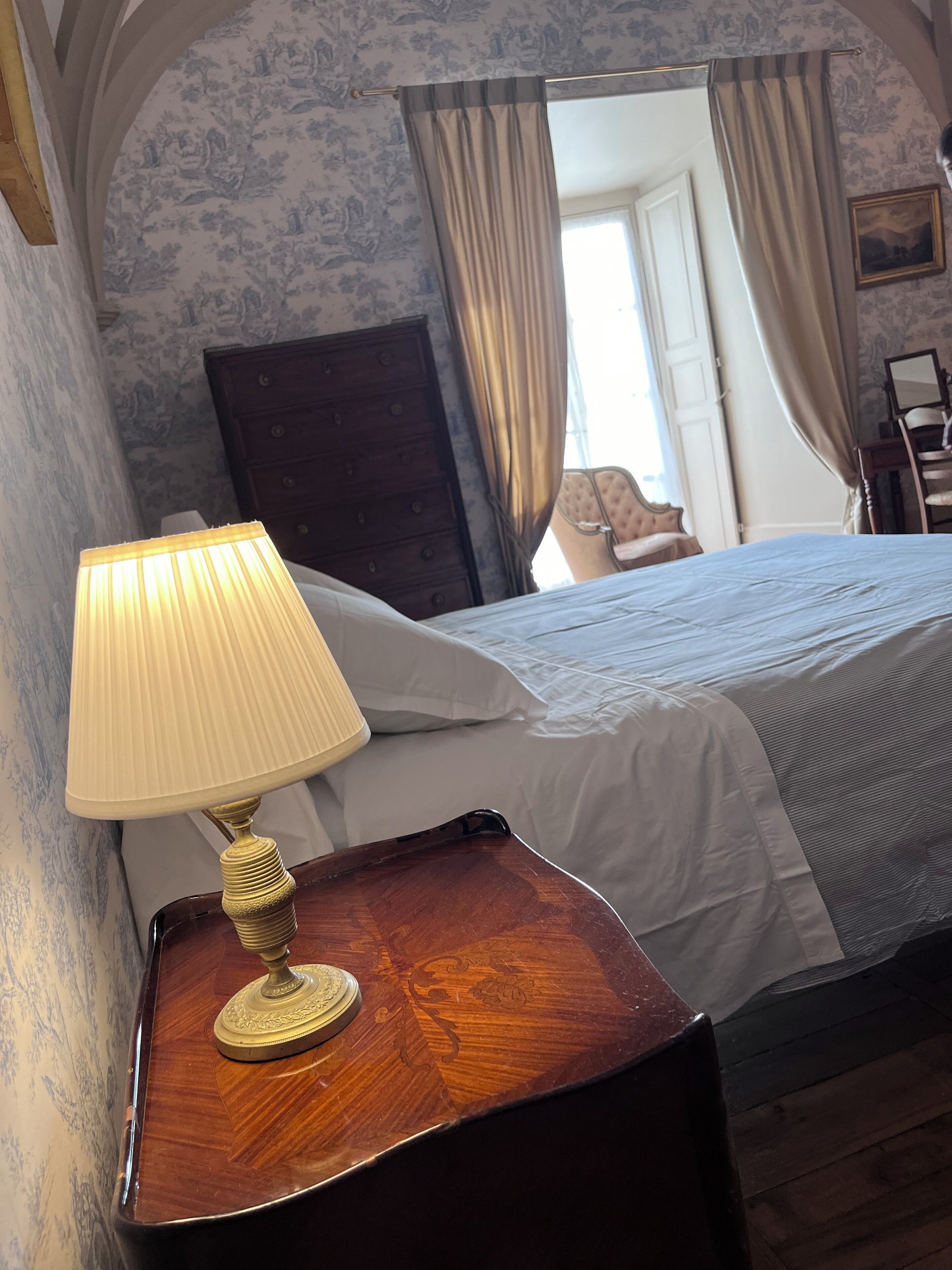 A bedroom with a wooden nightstand, a lamp, a bed with white linens, a dark dresser, and a mirror on the wall.