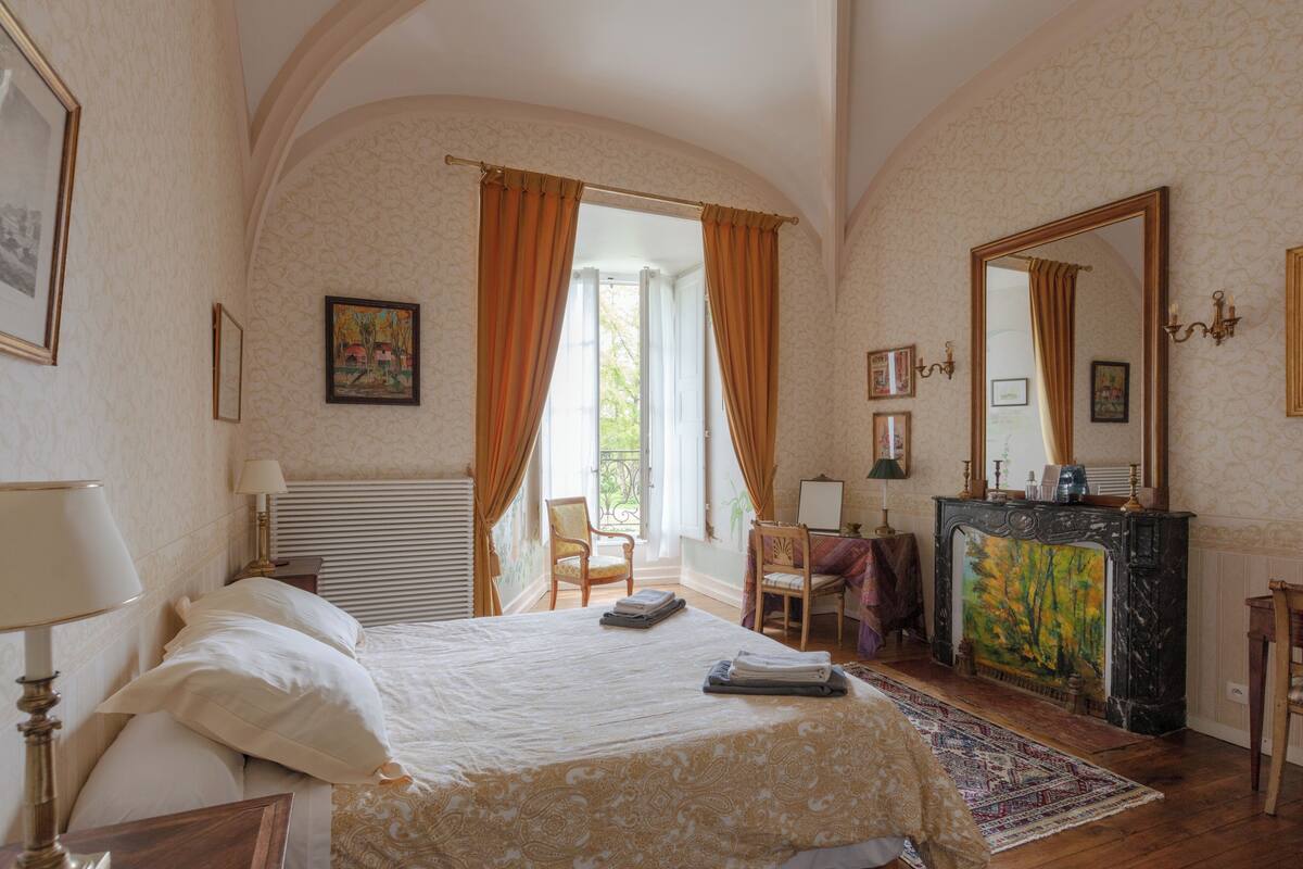 Quarto com cama, poltrona, escrivaninha, lareira e um espelho grande.