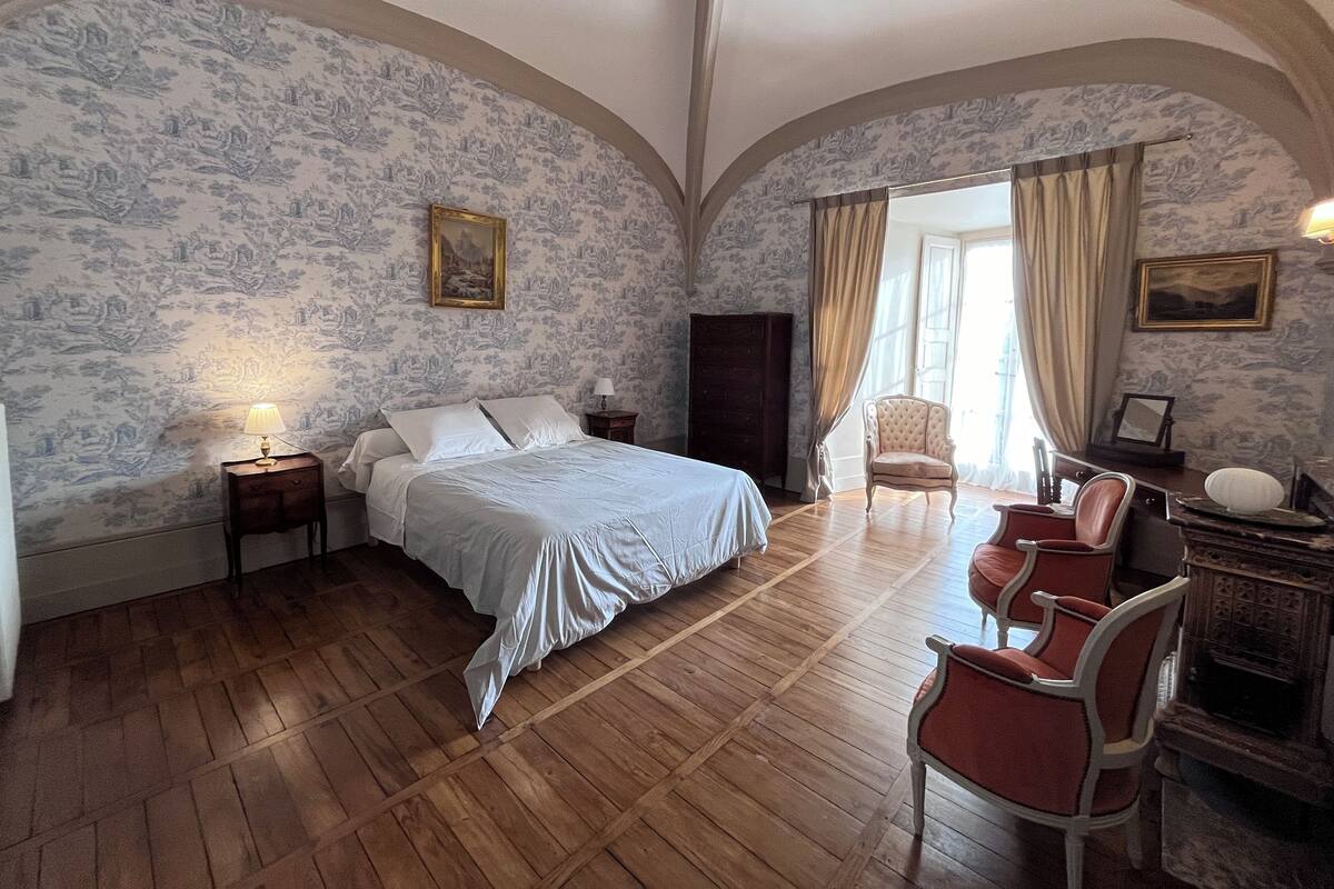 Quarto com uma cama grande, piso de madeira, uma cadeira, uma escrivaninha e uma lareira.