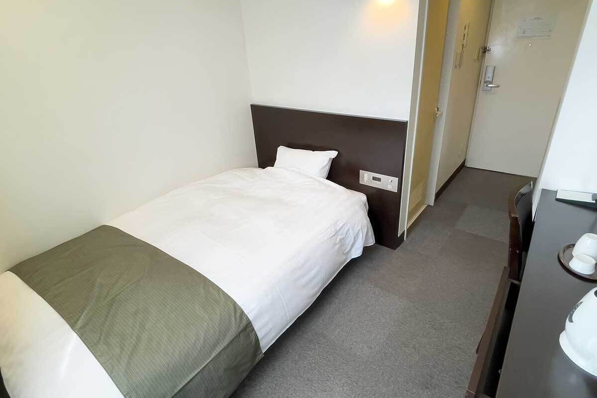 Habitación de hotel pequeña y ordenada, con una cama individual, una mesita de noche, una silla y un escritorio con su respectiva silla.