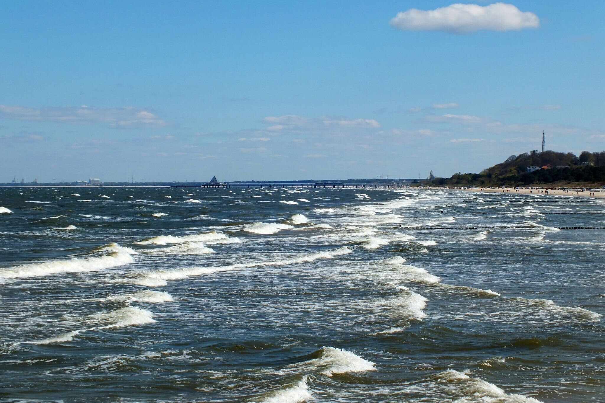 Blick auf die Küste mit Wellen, einem Steg und einem Strand voller Menschen.