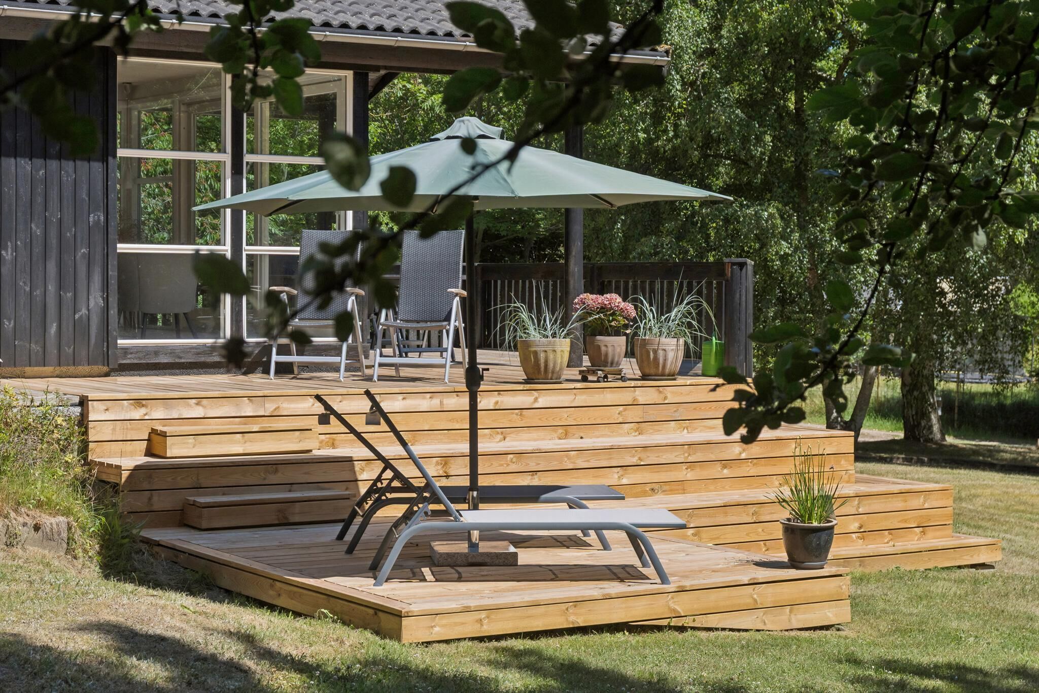 A wooden deck with a lounge chair and umbrella, surrounded by potted plants and a covered seating area.