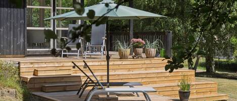 A wooden deck with a lounge chair and umbrella, surrounded by potted plants and a covered seating area.