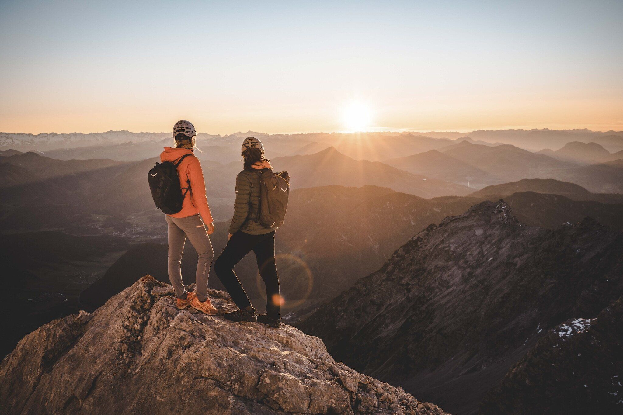 Deux personnes se tiennent debout au sommet d’un pic rocheux, avec une chaîne de montagnes en arrière-plan et le soleil couchant.