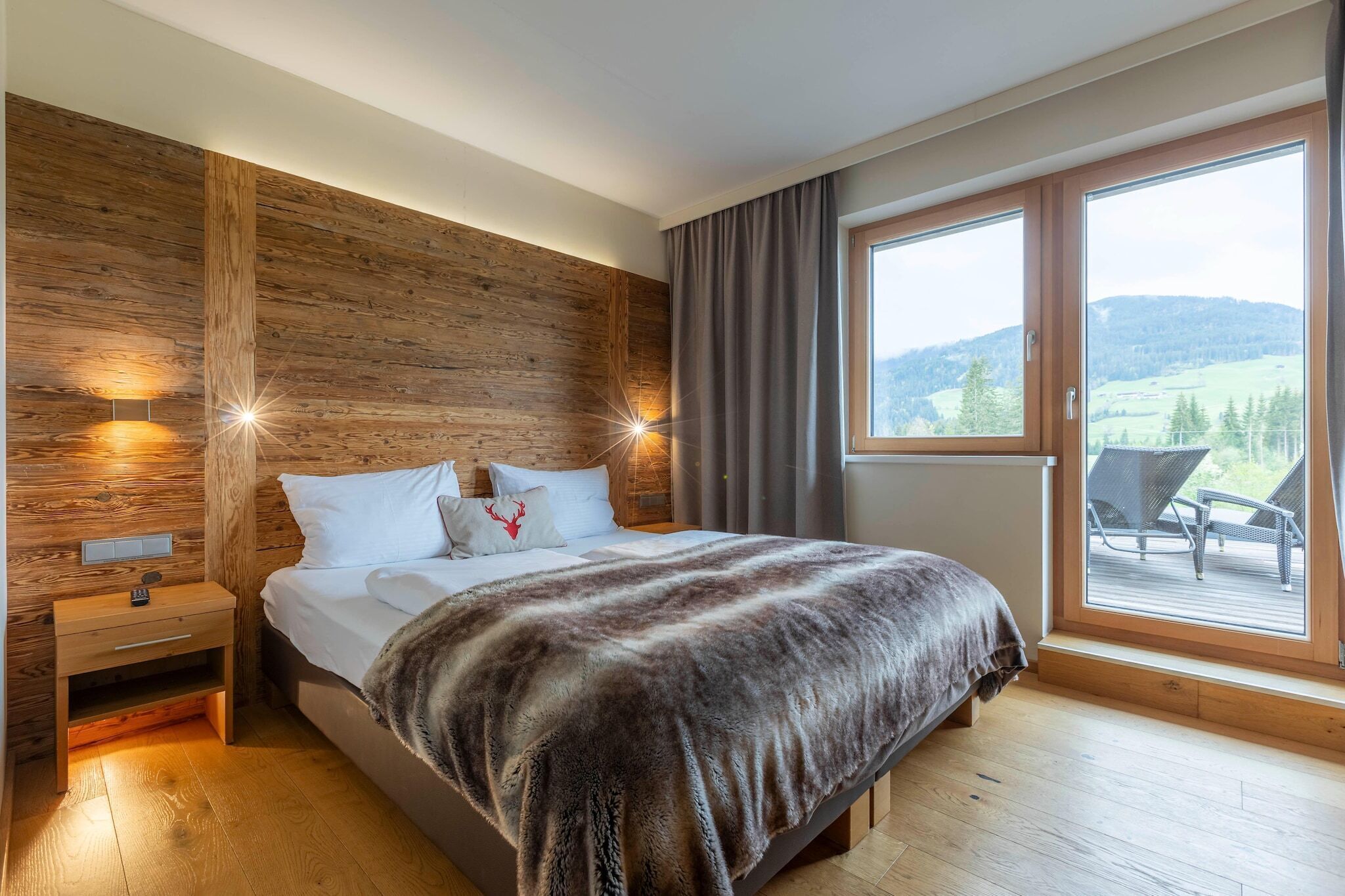 Quarto com parede de madeira, uma cama grande com manta de pele, mesa de cabeceira e vista para um deck e montanhas.
