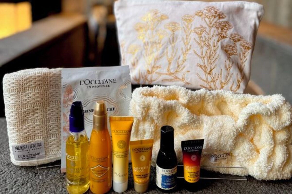 Una selección de productos L'OCCITANE, incluyendo botellas y una bolsa de tocador, dispuesta sobre una superficie texturizada con un cojín decorativo al fondo.