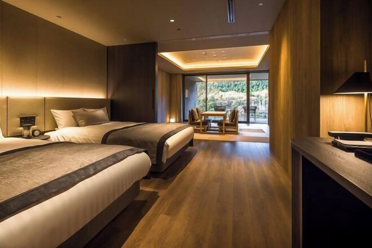 A modern hotel room with two beds, a dining area, and a view of greenery.