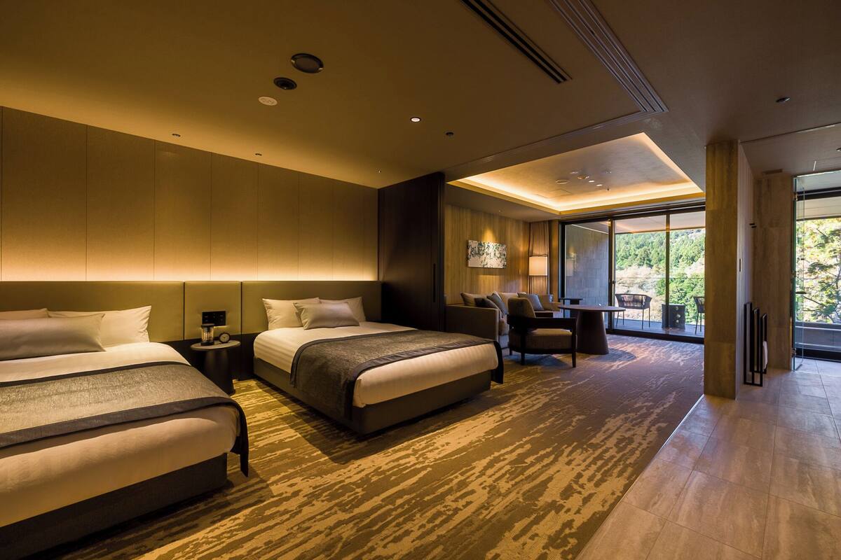 A modern hotel room with two beds, a seating area, and a large window offering a view of the outdoors.
