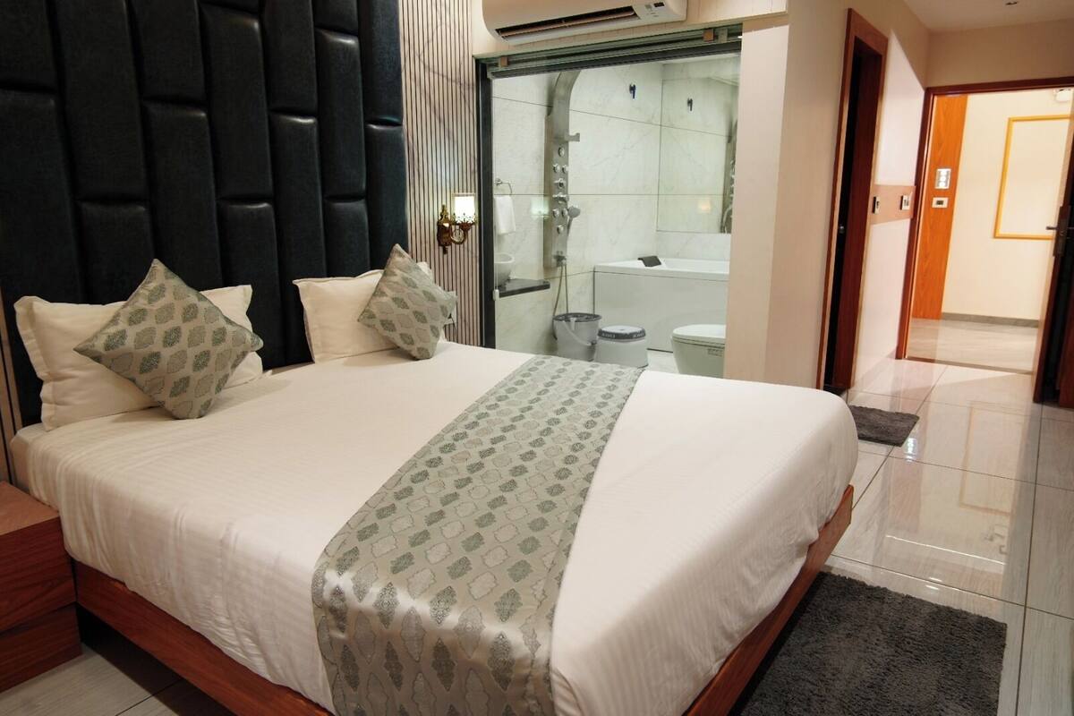 A hotel room with a large bed, a headboard, a bathroom with a glass door, and a wooden floor.