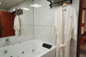 A bathroom with a marble wall, a bathtub, a wooden door, and a white robe hanging on a rack. - HOTEL BHAGYALAXMI LUXURY INN (Gandhinagar)