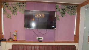 A modern living room with a sectional sofa, a round coffee table, a flat-screen TV mounted on a pink wall, and decorative plants. - HOTEL BHAGYALAXMI LUXURY INN (Gandhinagar)