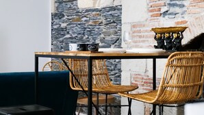 Una mesa con una cafetera, una cesta y un vaso. Hay sillas de mimbre y un sofá azul. El fondo muestra una pared de piedra y una pared de ladrillo.