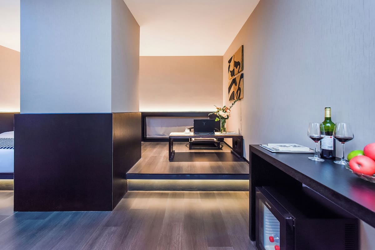Ein modernes Hotelzimmer mit dunklem Holzboden, einem Bett, einem Schreibtisch mit einem Laptop, einem kleinen Tisch mit Blumen in einer Vase und einer Minibar.