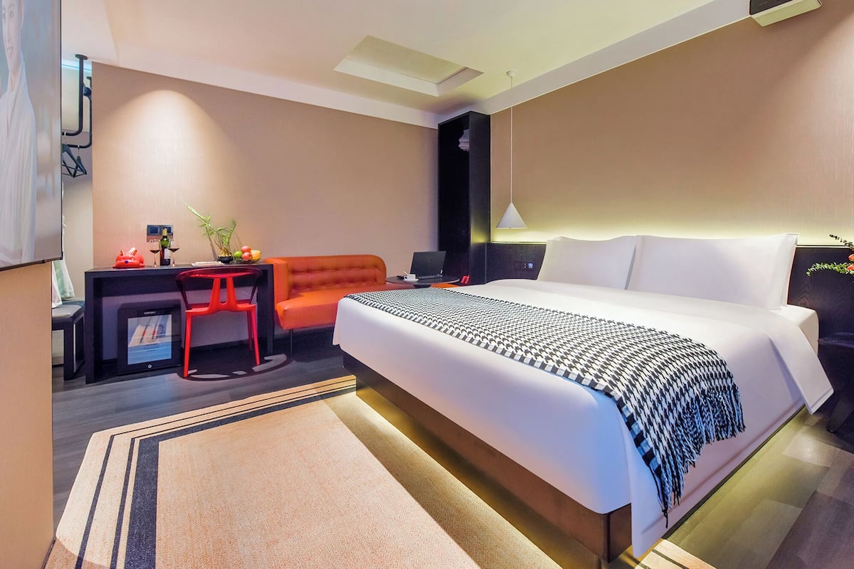 Ein modernes Hotelzimmer mit einem großen Bett, einem Schreibtisch mit Computer, einem roten Sessel und einem kleinen Tisch mit Pflanze und Obst.